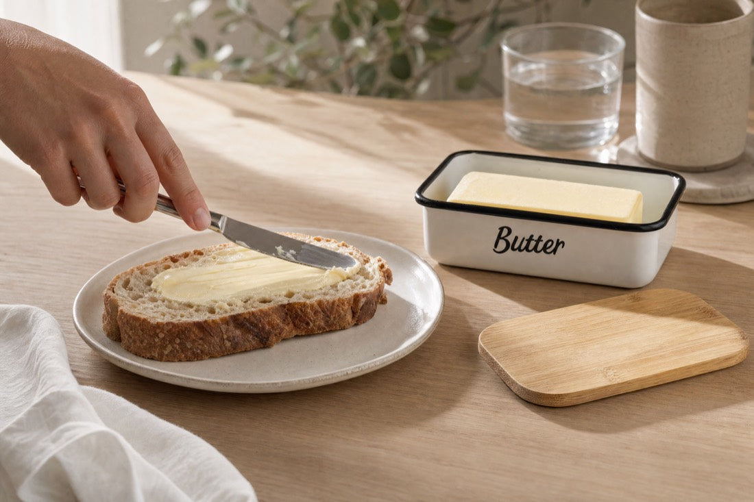 A hand spreading soft butter smoothly across a slice of sourdough, the enamel butter dish open beside it