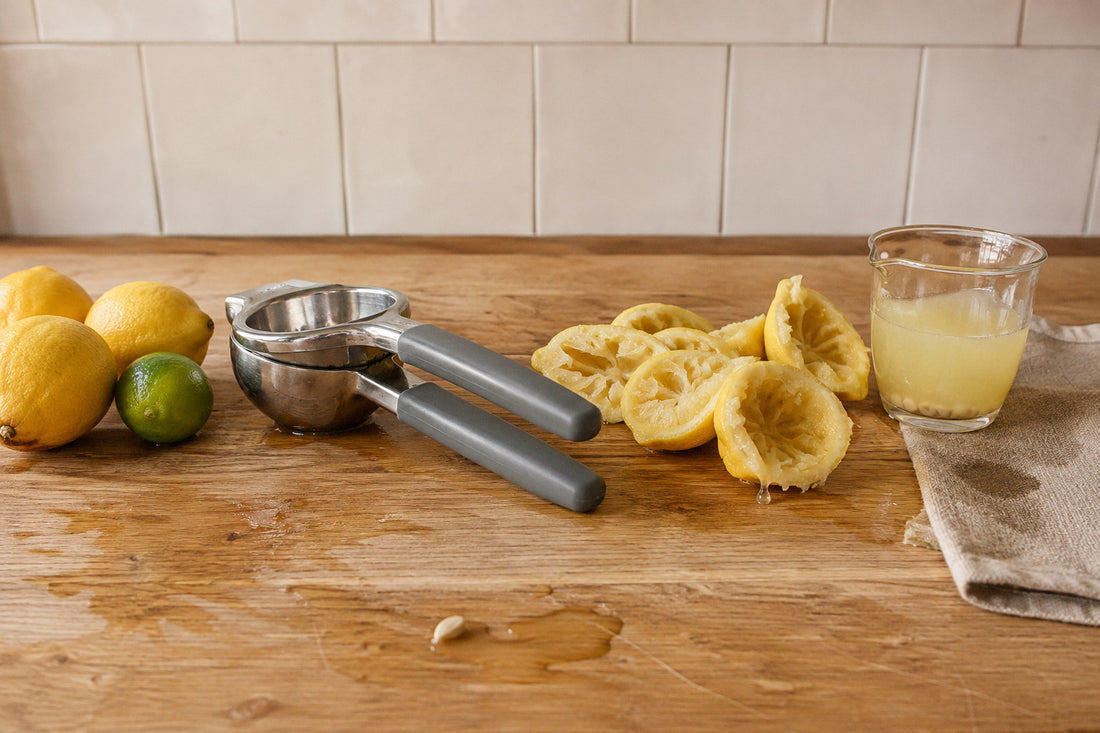 Stainless steel lemon squeezer lying on an oak board after pressing, spent lemon halves turned inside out beside it and a jug of juice