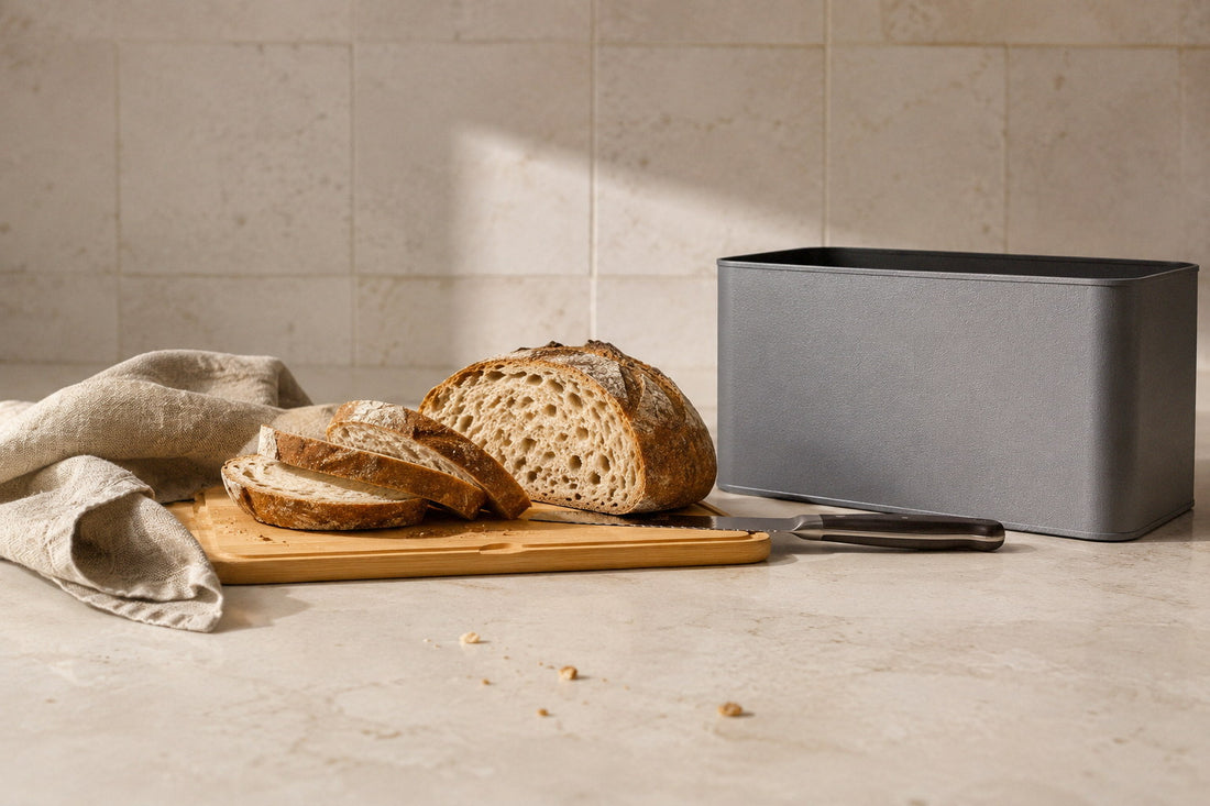 Grey bread box on a stone counter with its bamboo lid used as a board, a half cut sourdough loaf and a bread knife on it