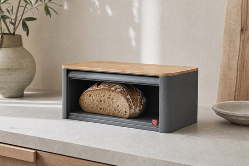 The steel bread box with its roll top open and a cut sourdough loaf inside, bamboo lid on top