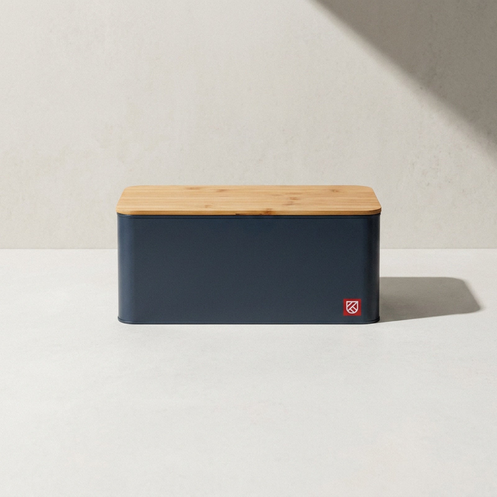 Kensington London charcoal steel bread box with bamboo board lid and red shield badge on pale limestone